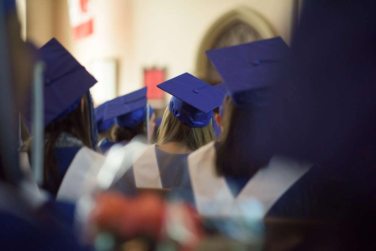 Editorial Photography For Cdi College - 2 Image Asset Students donning caps and gowns, sit waiting for their names to be called during the graduation ceremony. The photograph is captured at eye-level through rows of seated students, offering an interesting perspective and creating a sense of being imme…