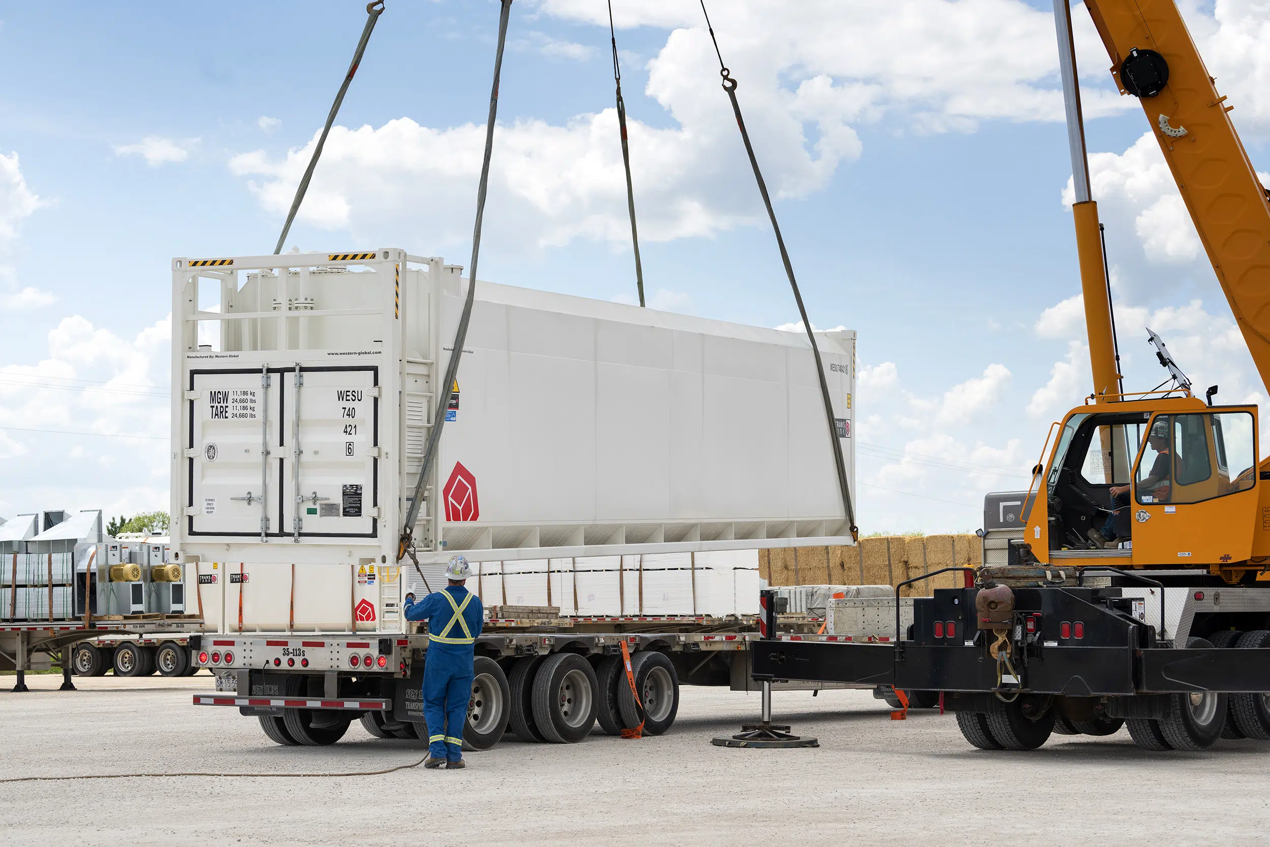 A crane lowers a very large fuel tank onto a semi trailer. A male worker in blue coveralls guides the operation.