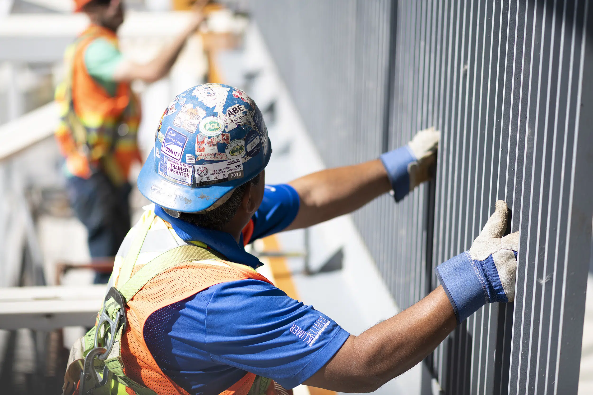 A worker in the foreground holds up a steel guard to be fastened by a worker in the distance. They are working at heights.